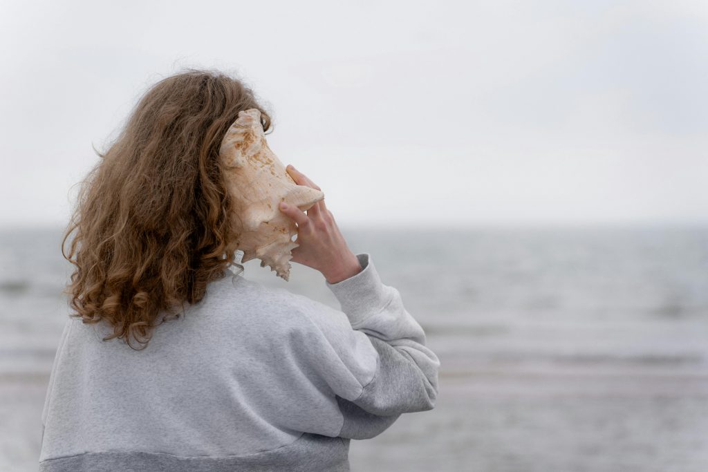 Woman holding a conch shell to her ear while standing on a quiet beach, listening to the sound inside the shell.